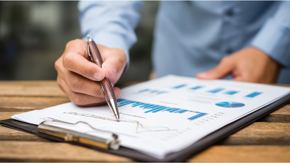 Close-up of a hand pointing at a clipboard with financial graphs and charts, indicating performance trends in a business report.