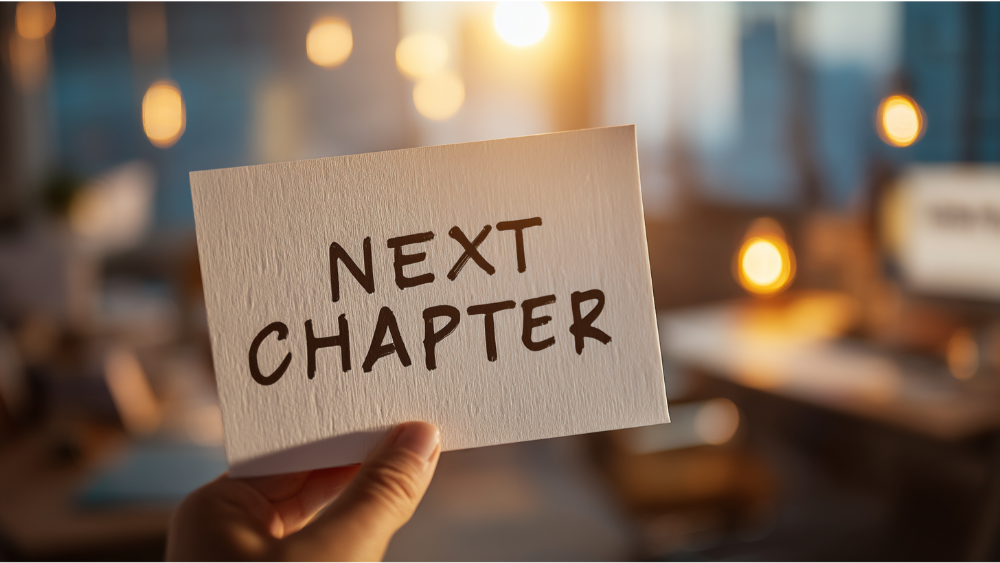 Close-up of a hand holding a card that reads ‘Next Chapter’ with blurred office lights in the background