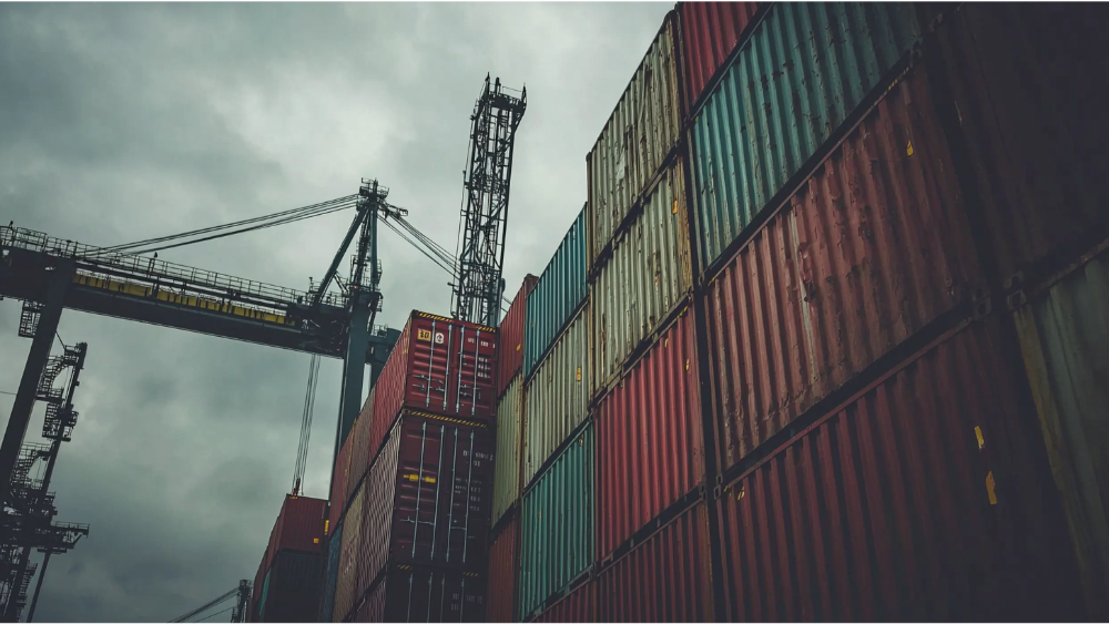 Stacked cargo containers at an industrial shipping port, with cranes and overcast skies in the background.