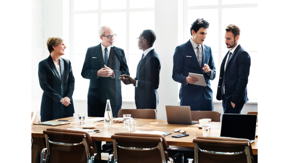 Five business people in formal attire engaging in conversation around a conference table with laptops and water glasses