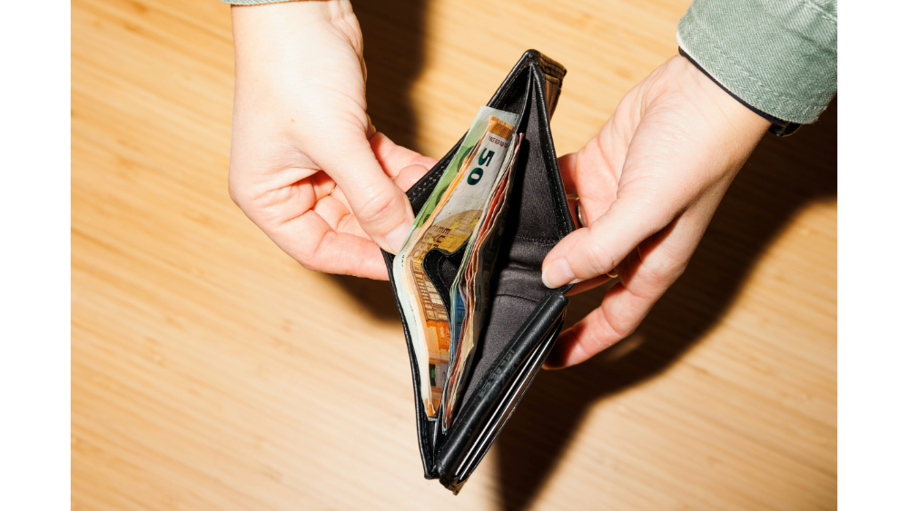 Close-up of hands holding an open wallet containing various currency bills, displayed over a wooden surface.