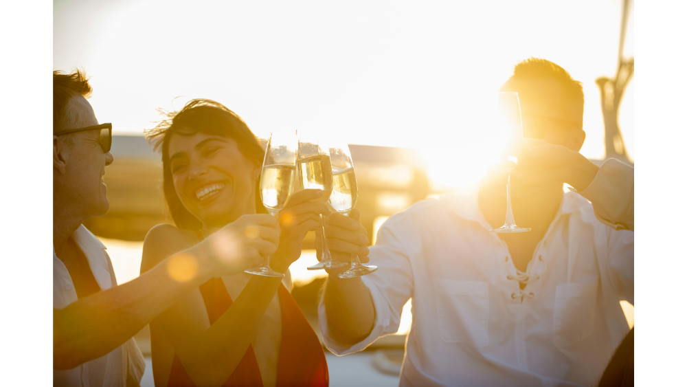 Group of people clinking glasses and laughing on a boat during golden hour, with warm sunlight creating a joyful mood.