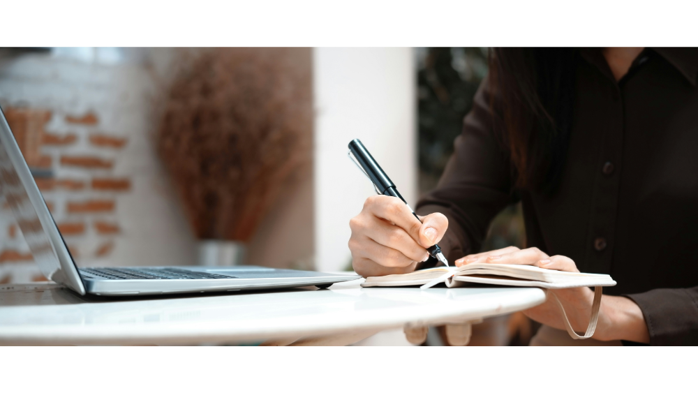 Woman writing in a notebook with a pen while working at a laptop on a round table, in a casual indoor setting.