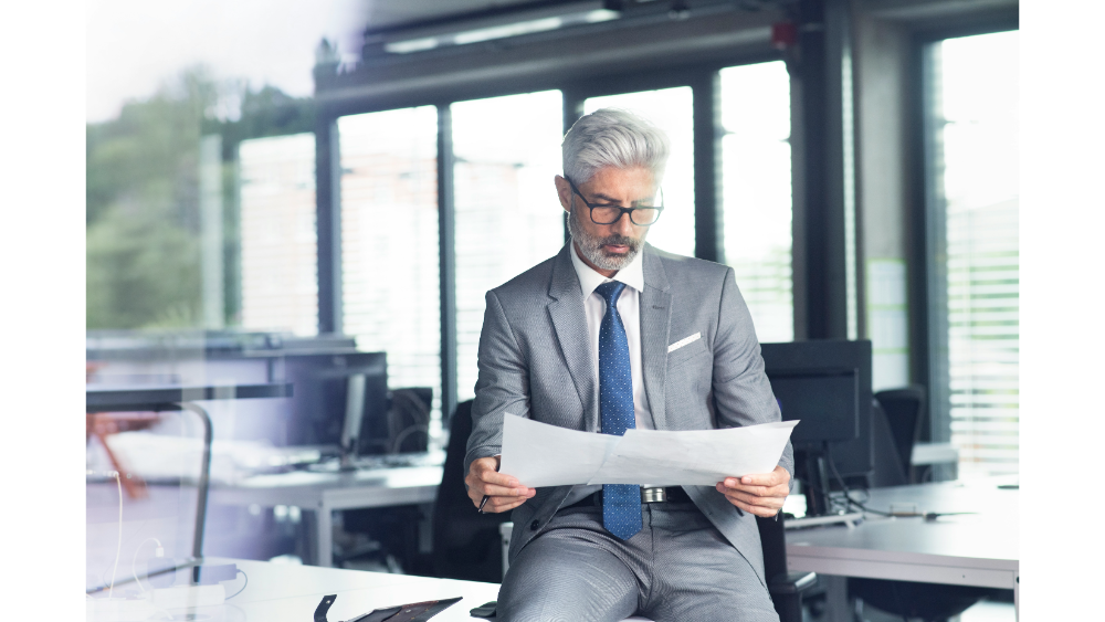 Mature businessman in a gray suit sitting on a desk in a modern office, reviewing financial documents with a focused expression.