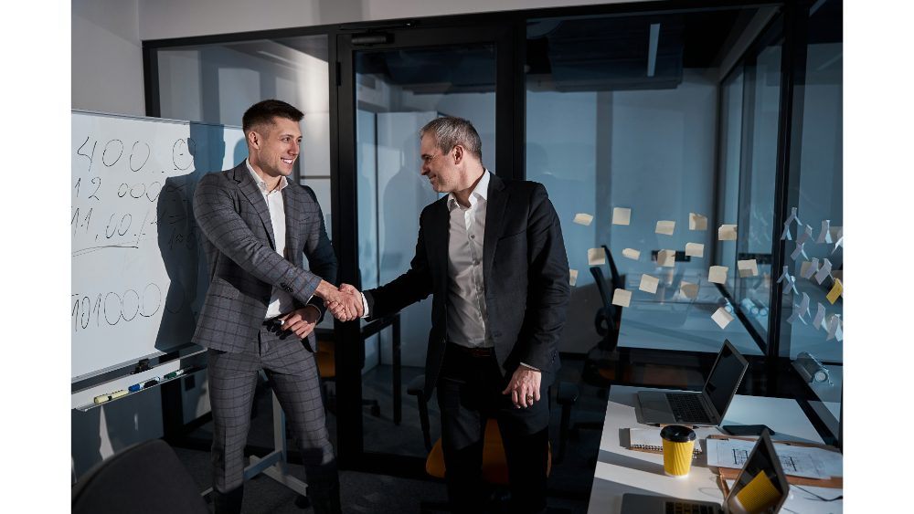 Two professionally dressed men shaking hands in a modern office, standing near a whiteboard and glass wall covered in sticky notes