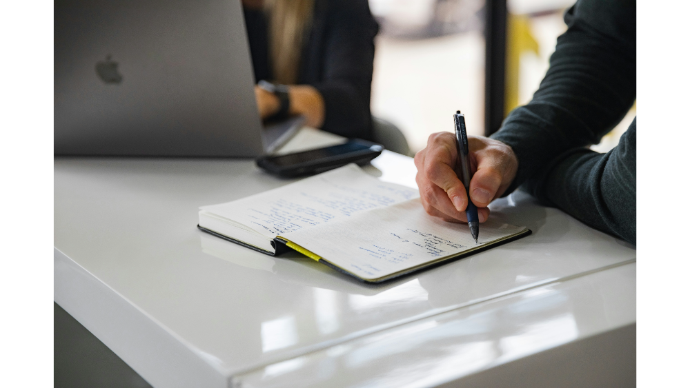 Close-up of a person writing notes in a notebook next to a laptop and smartphone on a white desk