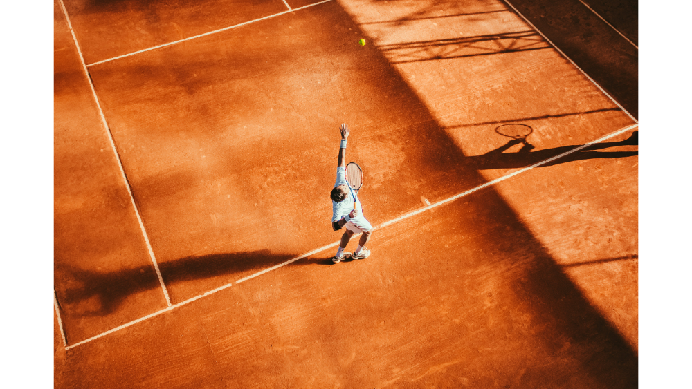 Aerial view of a tennis player serving on a clay court, with shadows and lines creating a dynamic visual composition.