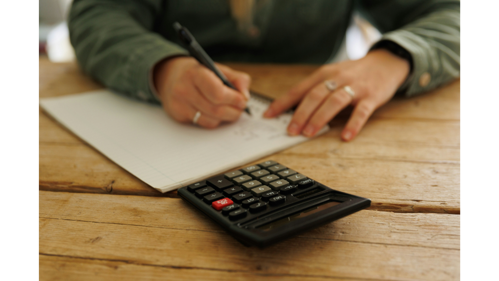 Close-up of someone writing in a notebook with a calculator nearby on a rustic wooden table, representing personal budgeting or financial planning.
