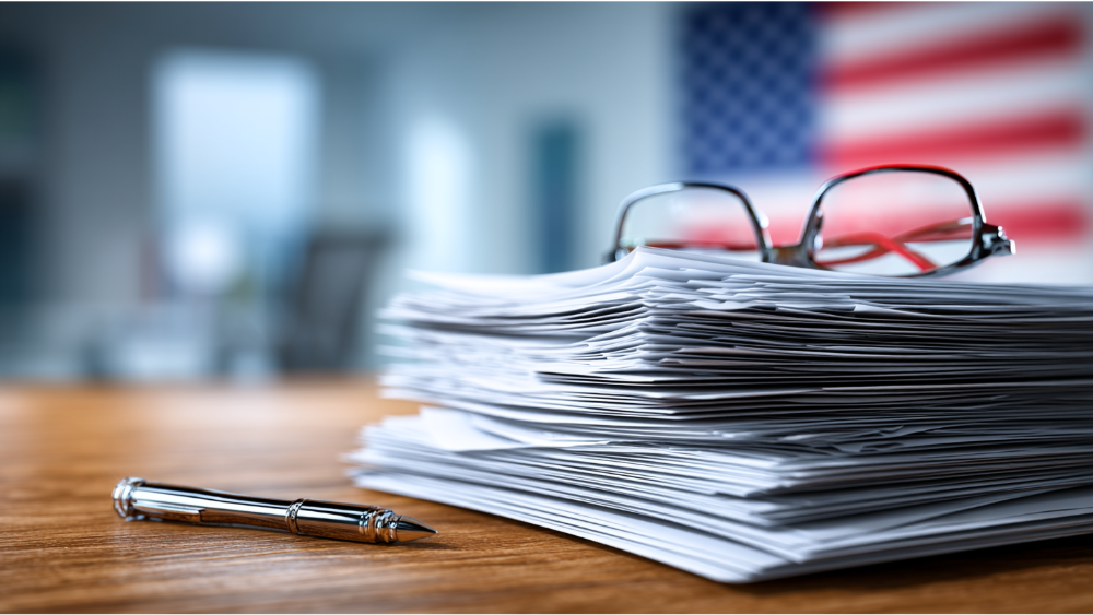 Tall stack of paperwork with a pen and reading glasses resting on top, with an American flag blurred in the background.