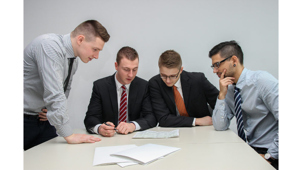 Group of four business professionals in suits gathered around a table, discussing paperwork in a meeting room.