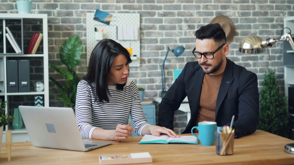 Two coworkers sitting at a desk in a modern office, reviewing documents together with a laptop and coffee mug nearby.